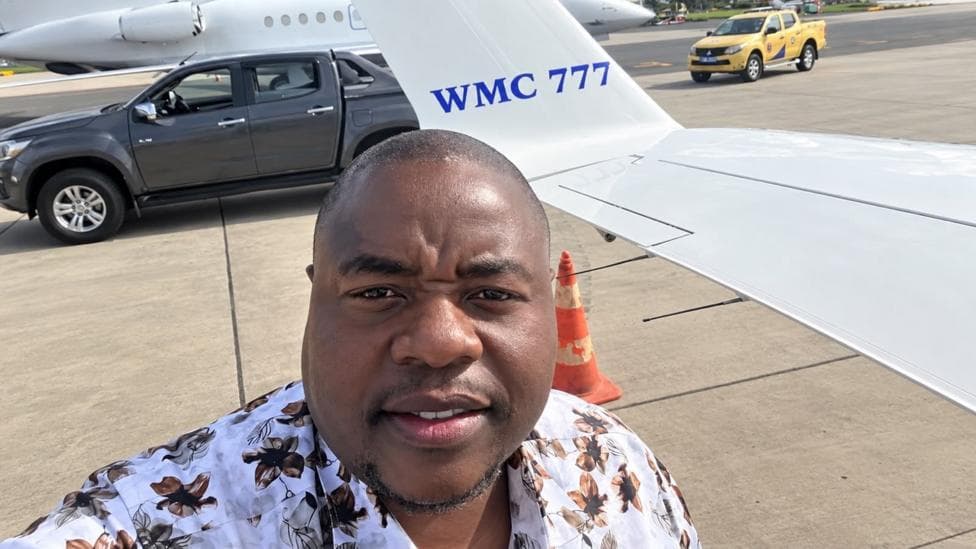 Wicknell Chivayo taking a selfie on an airport tarmac beside a small white aircraft marked “WMC 777.” A dark pick-up truck, a yellow airport vehicle and an orange traffic cone are visible behind him
