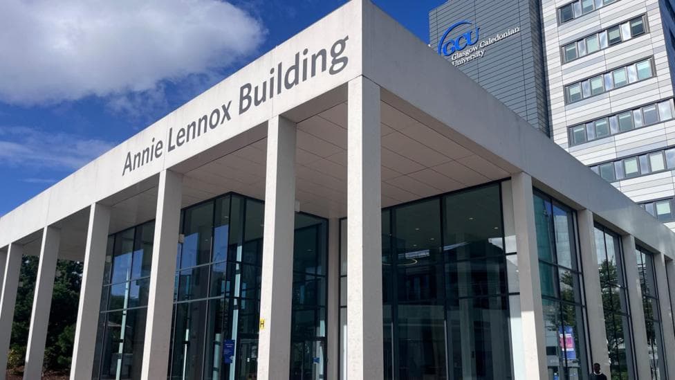 Exterior of Glasgow Caledonian University's Annie Lennox Building, showing the modern glass-fronted entrance and white-columned facade beneath a blue sky. The GCU logo is visible on the adjoining tower.