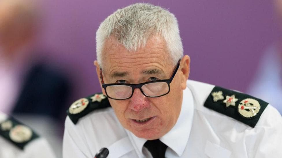 Jon Boutcher is looking over the top of his glasses which have a black frame. He has short, white hair. He is wearing a shirt with epaulettes on the shoulders and a black tie.