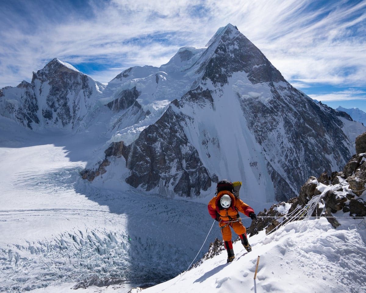 A mountaineer in an orange suit climbs a roped snowy ridge on K2, towering peaks behind