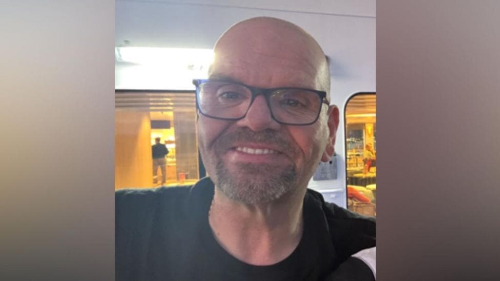 A man is smiling into the camera. He wears black glasses and has a black top on.