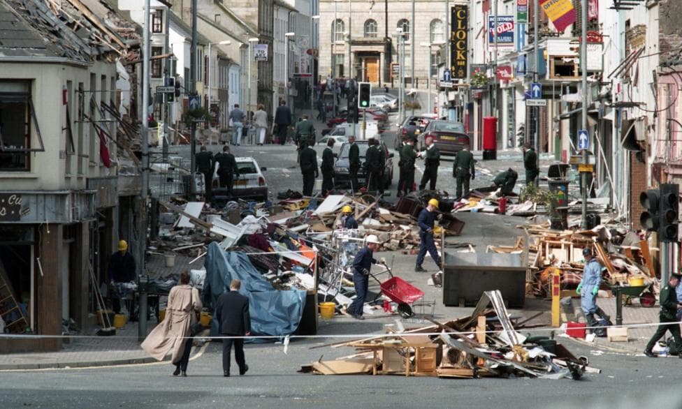 Police tape is cordoning off a street after a bomb. Debris is lying all over the street. People wearing hard hats are working.