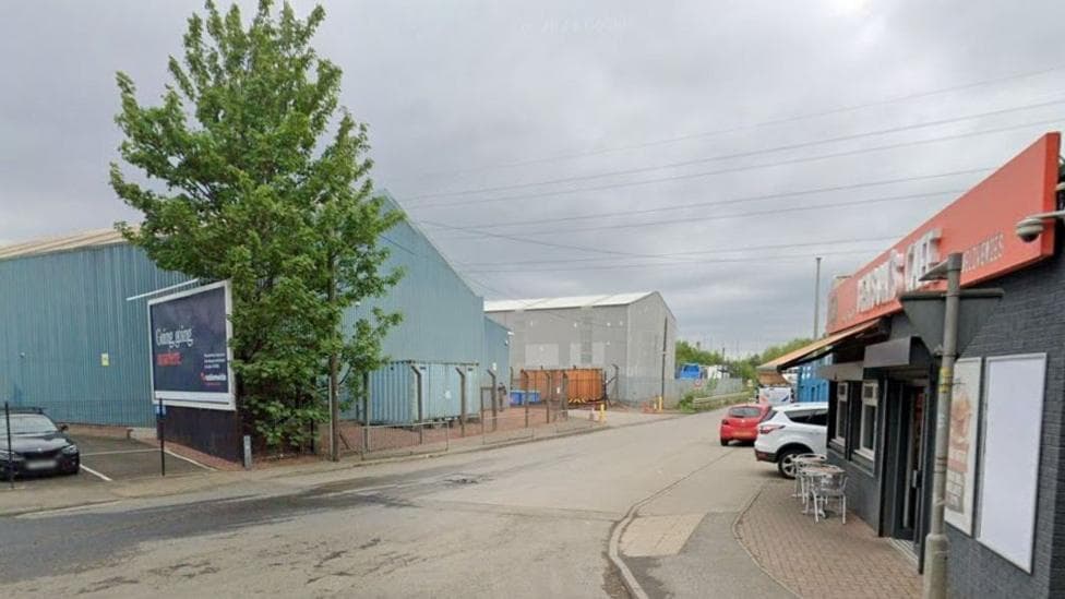 A road leading into an business park, with a cafe on one side of the street and a tree and car park on the other side. Cars can be seen parked further down the street as well.