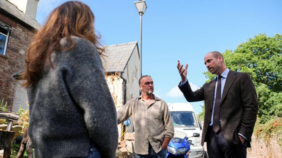 A back of a woman with long brown hair looking at Prince William, who is wearing a suit. A man in a casual shirt is also looking at William. They're stood outside a house.