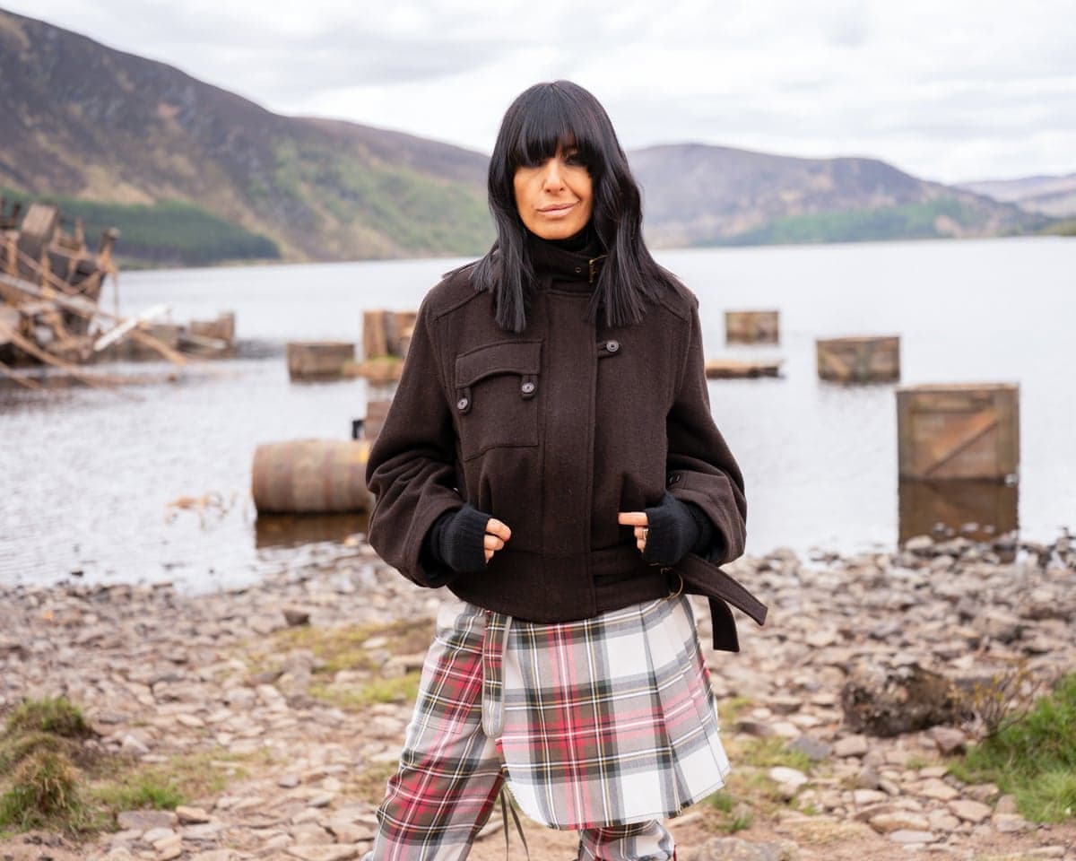 Claudia Winkleman stands in a dark jacket and tartan skirt by a lake with mountains behind