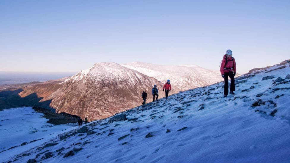 Hikers walk in a line along snow-covered mountain slope speckled with exposed rock. With the first in the group you can see their white helmet and they are carrying a backpack. Snow-covered peaks and valleys stretch into the distance beneath a clear blue sky.