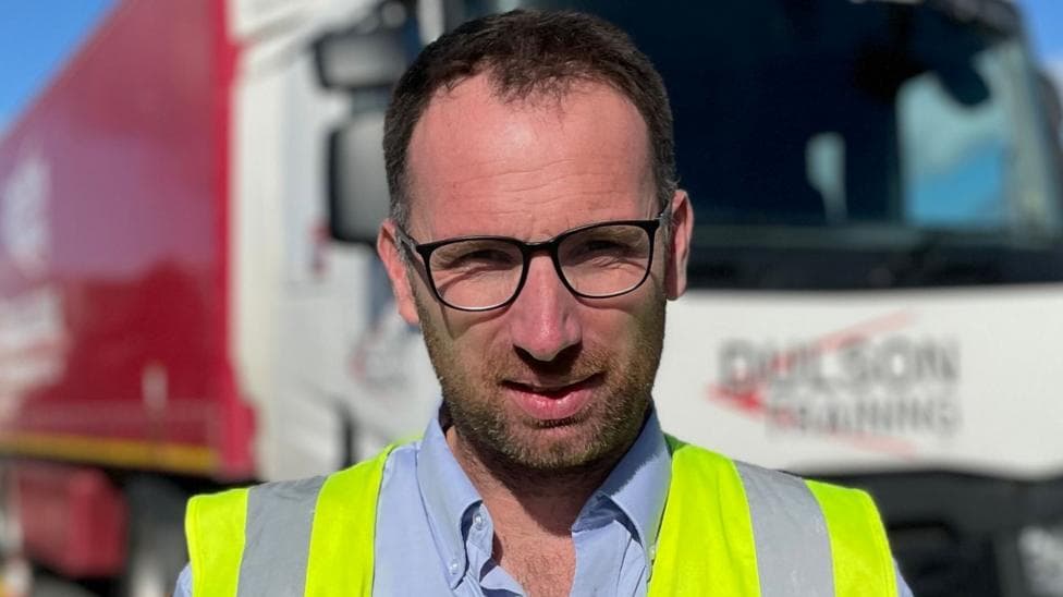 A dark-haired man wearing a blue short and high vis vest, standing in front of a white lorry with a red trailer. "Dulson Training" can be read on the front of the lorry.