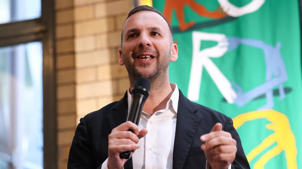 Green Party of England and Wales leader Zack Polanski wears a black blazer and white shirt. He holds a microphone to his mouth as he speaks.