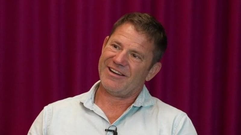 Steve Backshall is seated indoors in an upholstered chair during an interview. He is wearing a light-coloured buttoned shirt with a clip-on microphone attached. A floor lamp with two circular lights stands to one side, while deep red curtains form the background.