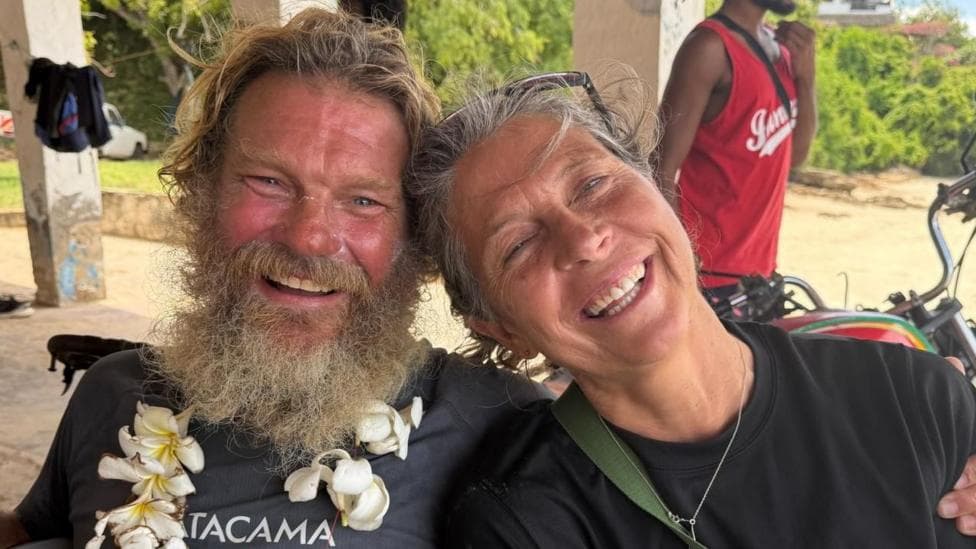 Dave Dinger Bell and his partner Georgie smile for the camera. Bell has a long scruffy beard and blond hair. Georgie has tied back hair.