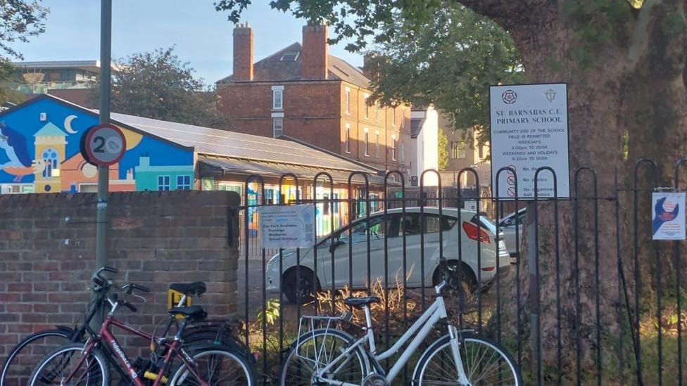 Outside St Barnabas CE Primary. Two bikes are parked against the gates. There is a poster for the repair shop and information about the school. It is a sunny day.