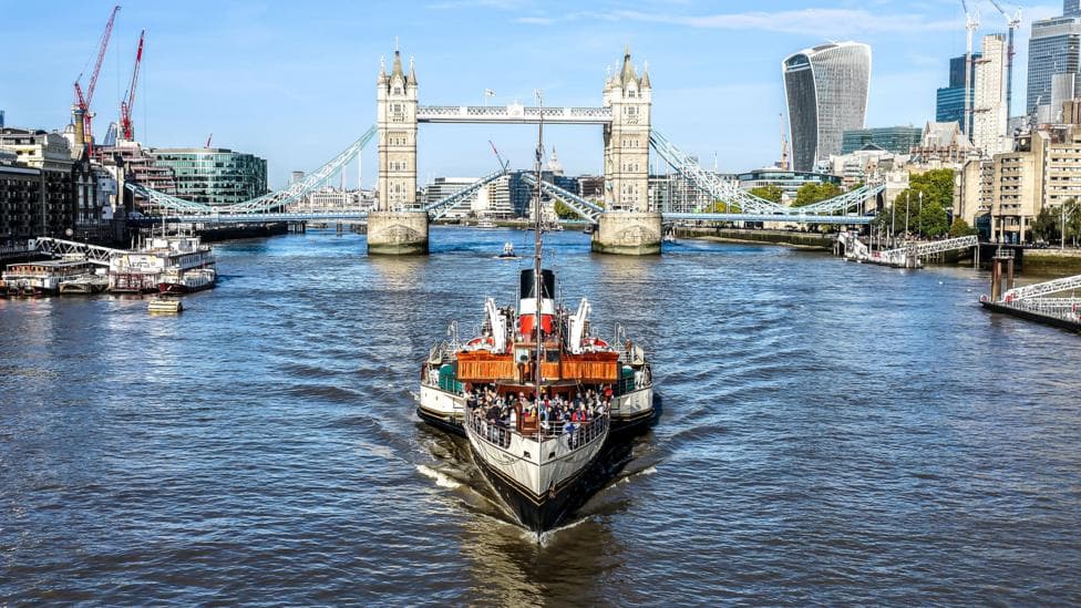 The paddle steamer Waverley sailing along the River Thames towards Tower Bridge in London, with passengers on deck and the city skyline under a blue sky in the background.
