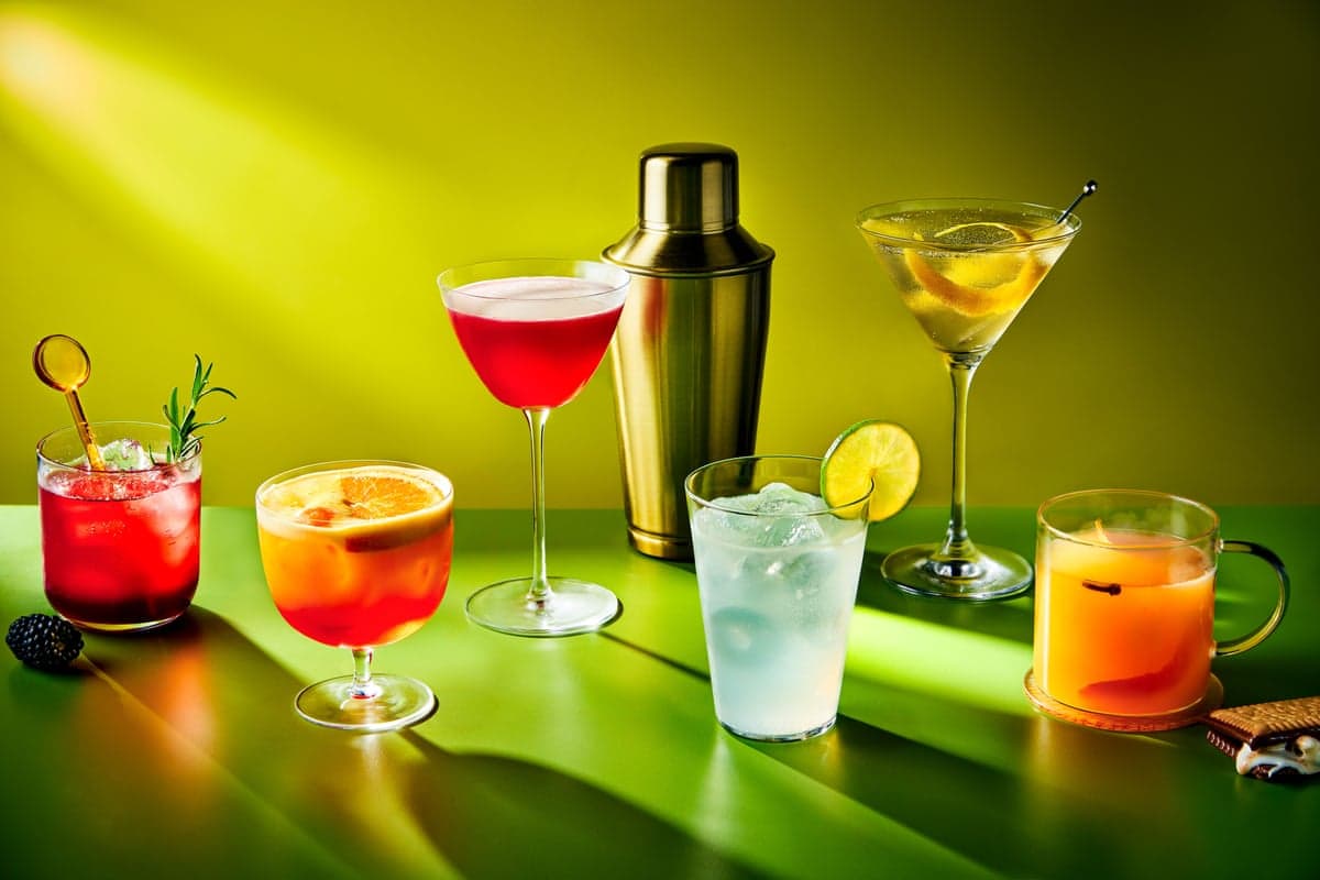 Six colorful cocktails and a metal shaker are arranged on a green table with green lighting