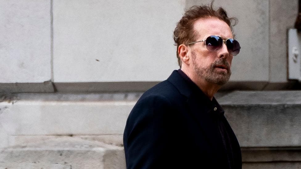 A bearded man wearing a blue jacket and sunglasses walks past a building.