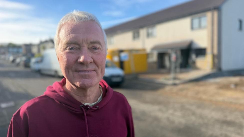 A man with short grey hair wearing a burgundy hoody. He is standing in front of some newly built houses.