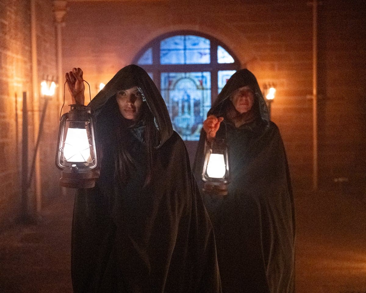 Maya Jama and Richard E Grant, hooded and cloaked, hold lanterns in a torch-lit corridor