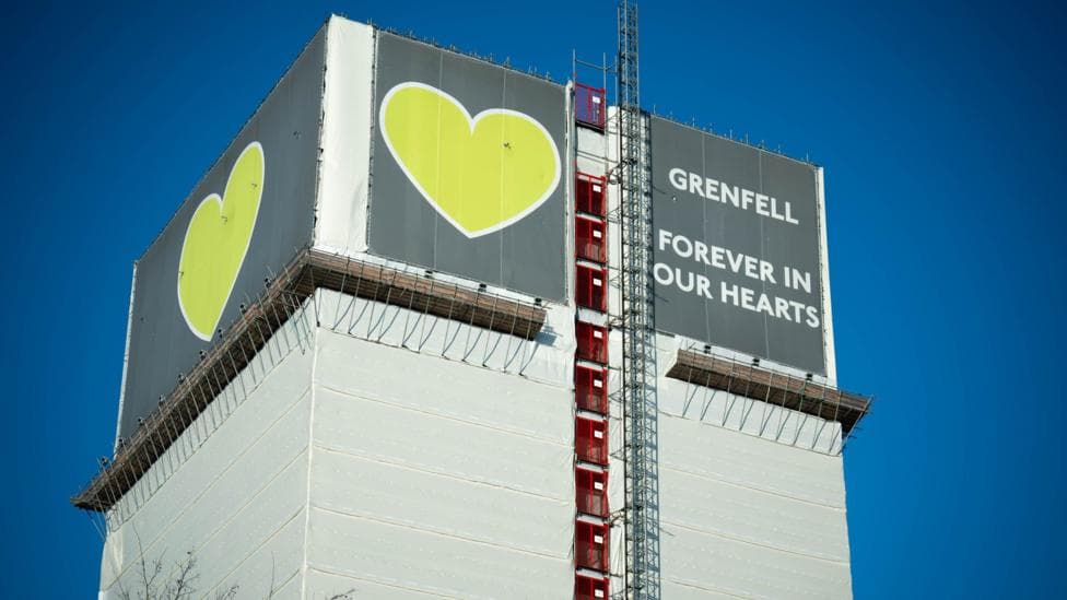 A tower block covered in white wrapping. There is a sign on top saying 'Grenfell - forever in our hearts'