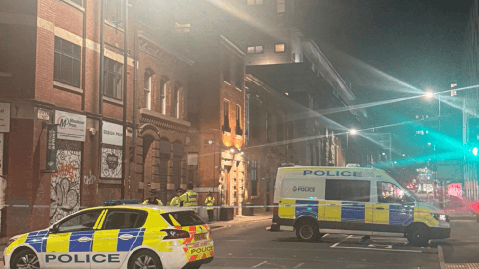 A police car and a van and several officers stand guard by a police cordon in a city-centre location.