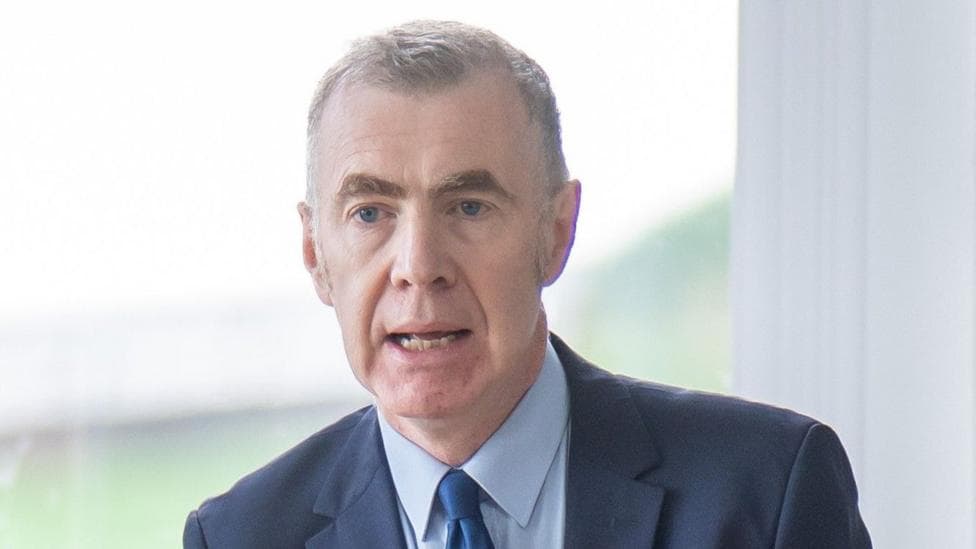 Adam Price speaking at an event in front of a window, wearing a blue suit, blue shirt and a blue tie.