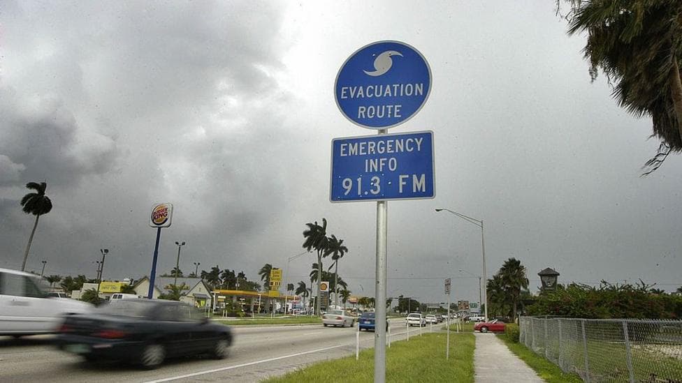 Photo shows a road with cars passing a blue sign that has a hurricane symbol and says 'evacuation route' 'emergency info 91.3FM'