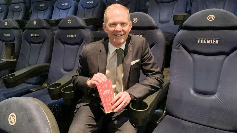 A man in a suit with thinning reddish/blond hair sits on a large cinema seat with empty seats all around him and a tub of popcorn in his hand