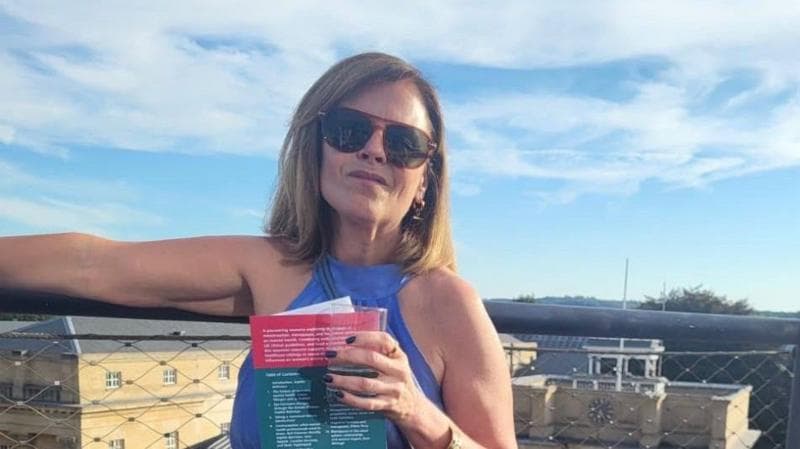 Sally williams is standing with one arm stretched out, holding a leaflet and a cup in the other. She is wearing a blue halter neck dress and sunglasses