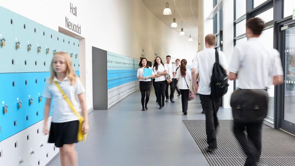 Pupils walking in school corridor in uniform of white polo shirt and black trousers or skirts. The building is modern and has blue and green lockers along one side of the wall and tall glass windows on the other side.