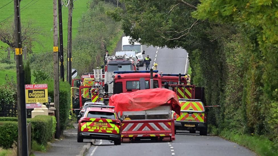 Emergency services including fire engines, fire cars and an ambulance parked close-by each other on a country road with trees and a footpath to the side.