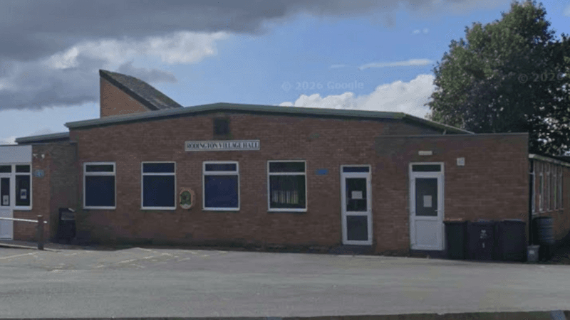 A single storey brick building with a sloped roof and a car park in the foreground