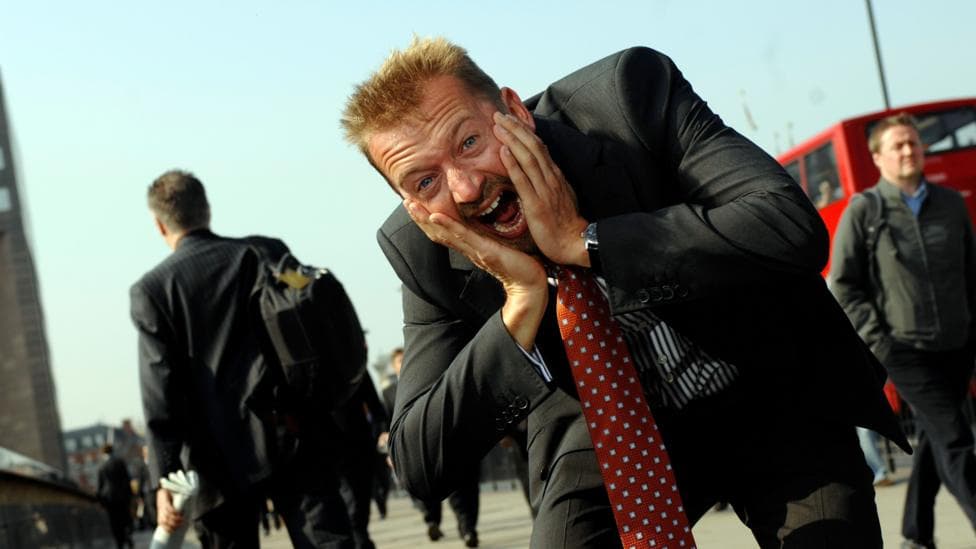 A man holding his face and screaming at the camera, He's wearing a suit and a red and blue polka-dot tie.