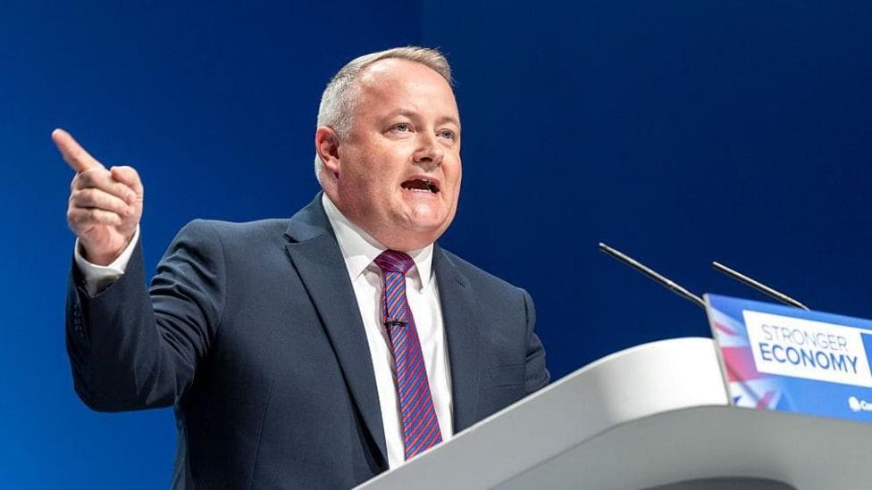 Darren Millar in navy suit and white shirt and tie at lectern talking and gesticulating right arm up and finger pointing
