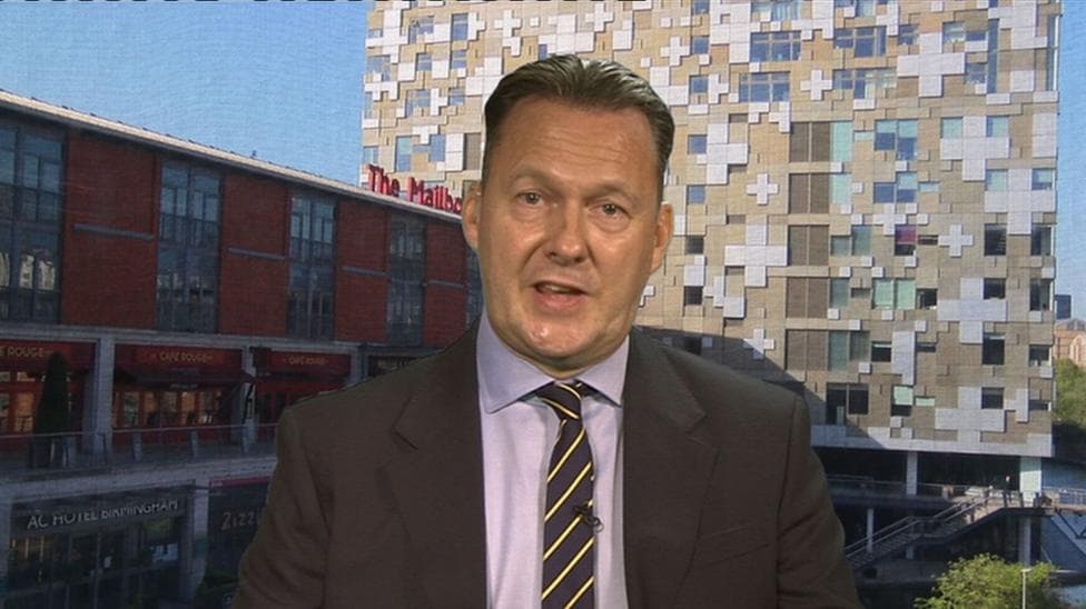 a man with slicked back brow hair is wearing a dark jacket, lilac shirt and blue and cream striped tie. He is being interviewed with a backdrop of images from a Birmingham street