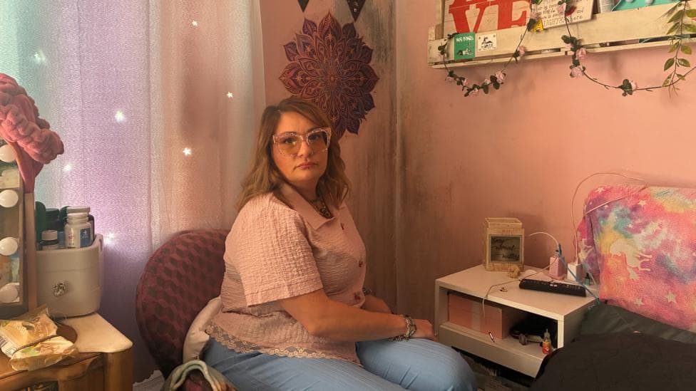 A woman with dark blonde hair with glasses sits in a child's bedroom. There is mould on the walls