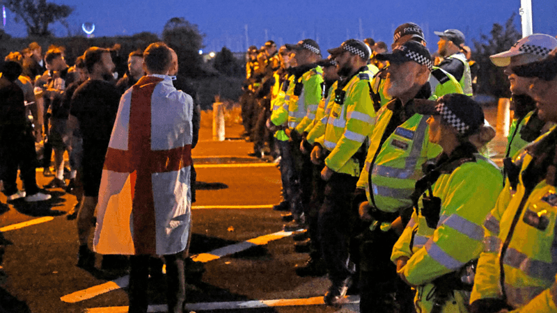 There is a line of police to the right of the image, facing towards the left. They're wearing yellow hi-vis jackets and black and white hats. On the left of the image are a handful of men, one wrapped in a red and white flag