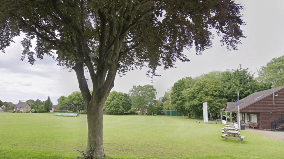 Parkland at Elmley Castle in Worcestershire. A large tree stands in the foreground with park benches and a brown club house on the edge of grass. Cricket covers and buildings can be seen in the background.