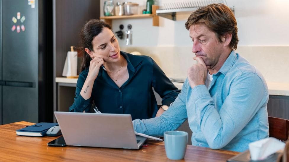 Man and woman sit at a kitchen table with a laptop and mug in front of them.