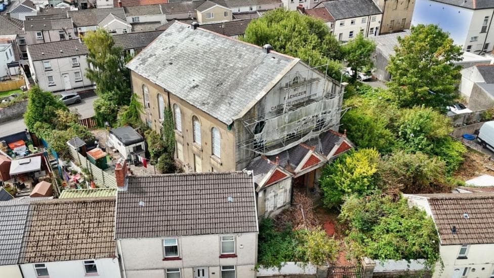 An aerial photo of a large stone chapel with a weathered slate roof, tall arched windows and scaffolding across the front. Overgrown trees and shrubs surround the building, with terraced houses, gardens and parked vehicles across the street behind the church. You can just make out Ebenezer written beneath the peak of the roof.
