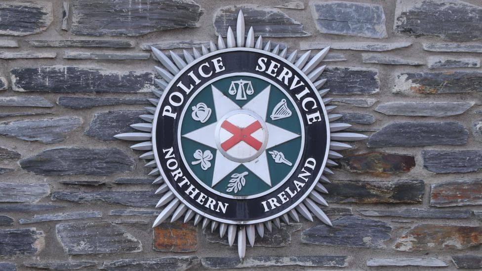 PSNI logo on a grey brick wall. The logo is red, black, white and green. It says Police Service Northern Ireland in capital letters.