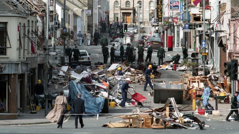 Police tape is cordoning off a street after a bomb. Debris is lying all over the street. People wearing hard hats are working.