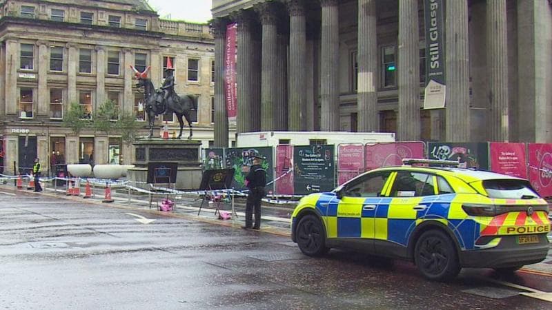 A police cordon surrounds an equestrian statue outside a columned city building in Glasgow, with a police vehicle parked nearby on a wet street.