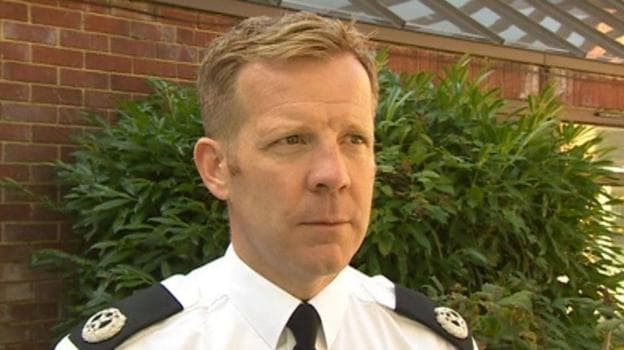 Scott Chilton has short brown hair and is in police uniform. He is standing in front of a bush and red brick building.