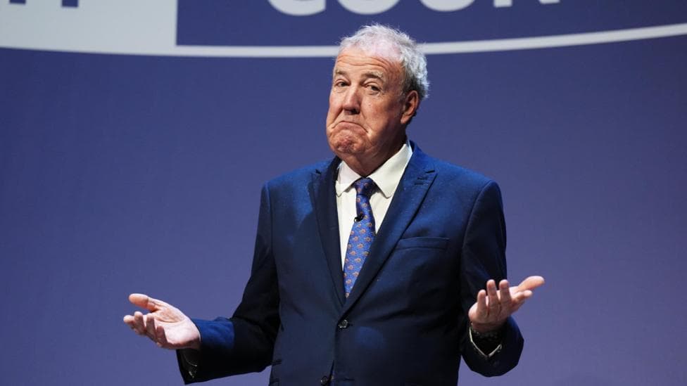 Jeremy Clarkson, in a suit and tie, shrugs his shoulders and holds up his palms on the Tory conference stage