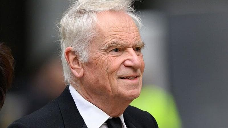 Jeffrey Archer is wearing a black suit and walking past the camera.