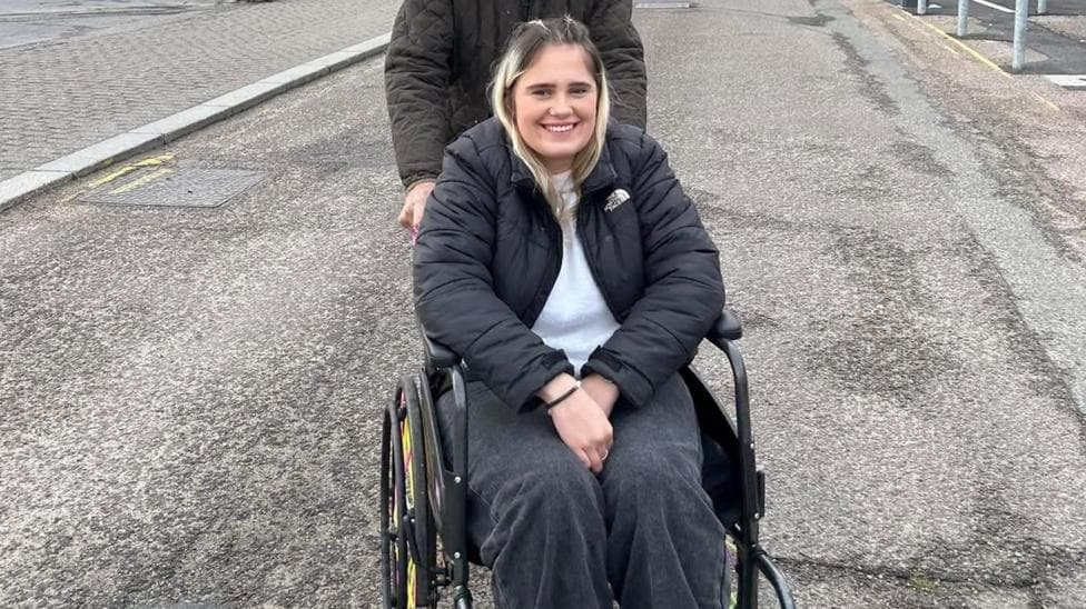 Phoebe Tesoriere is a wheelchair user. She has blonde hair and is wearing a black coat, white jumper and black jeans. She is smiling and looking at the camera. She is outside and being pushed in her wheelchair by a man behind her.