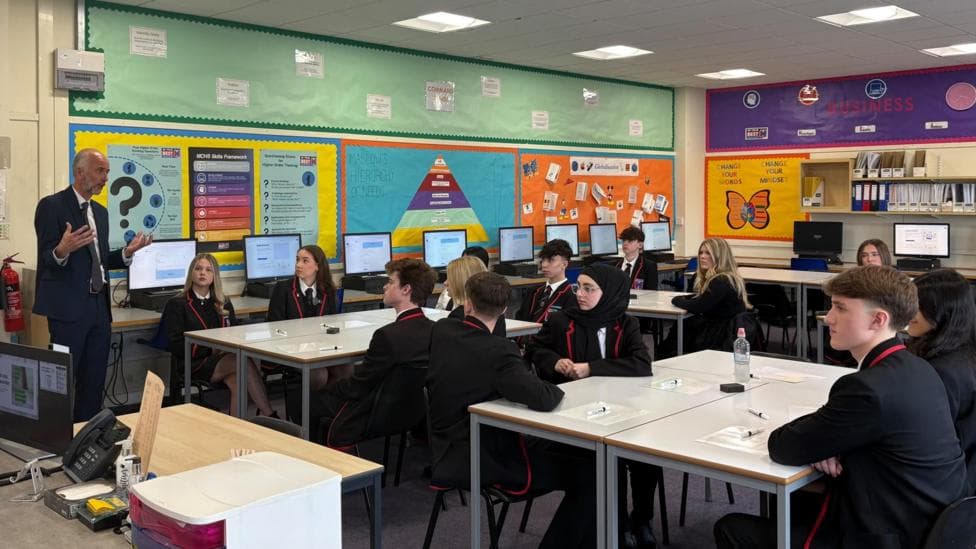 Classroom discussion with secondary school pupils in uniform seated around tables, listening to a speaker at the front of a computer-equipped business studies classroom.
