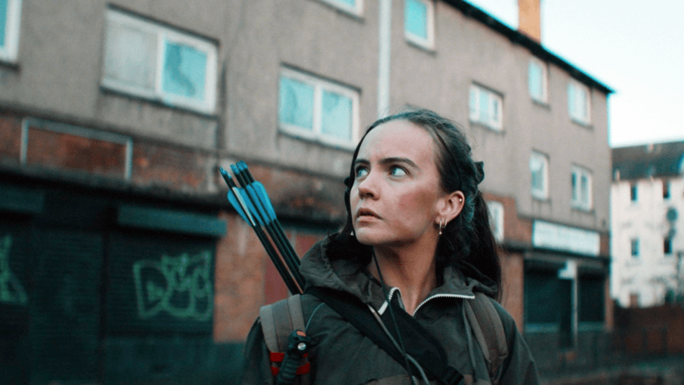 Jade McDonald is a young woman with dark hair, which is tied back. She is wearing dark clothing. Slung over her shoulder is a quiver with arrows in it. Behind Jade is part of the Clune Park Estate.