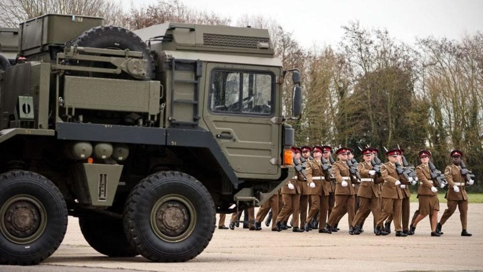 Troops march eyes right next to an Army vehicle.