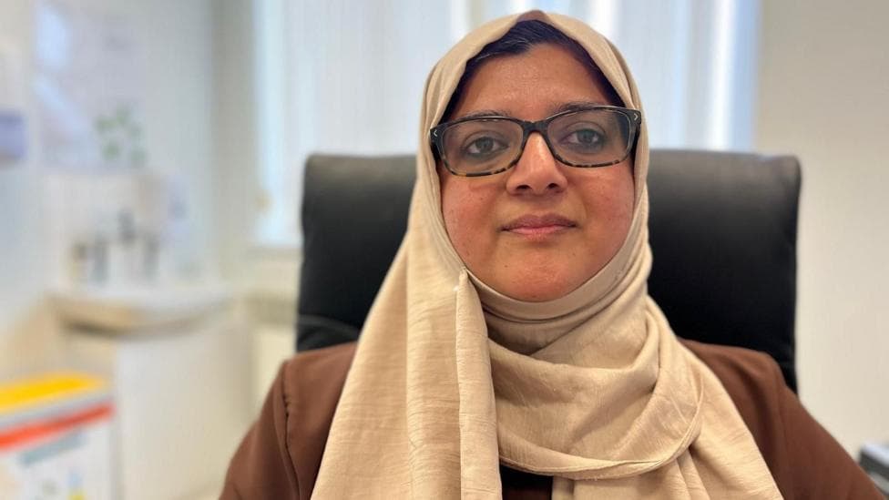 Dr Amara Naseem is sitting in a GP's surgery, she is wearing a cream-coloured headscarf and a light brown top. She has on rectangular tortoiseshell glasses and a medical bin can be seen over her right shoulder