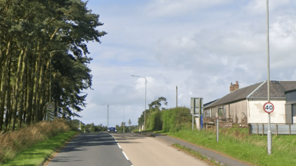 A stretch of road, with a 40 speed limit warning and a farm or house on one side of the road. On the other side of the road is grass and trees.