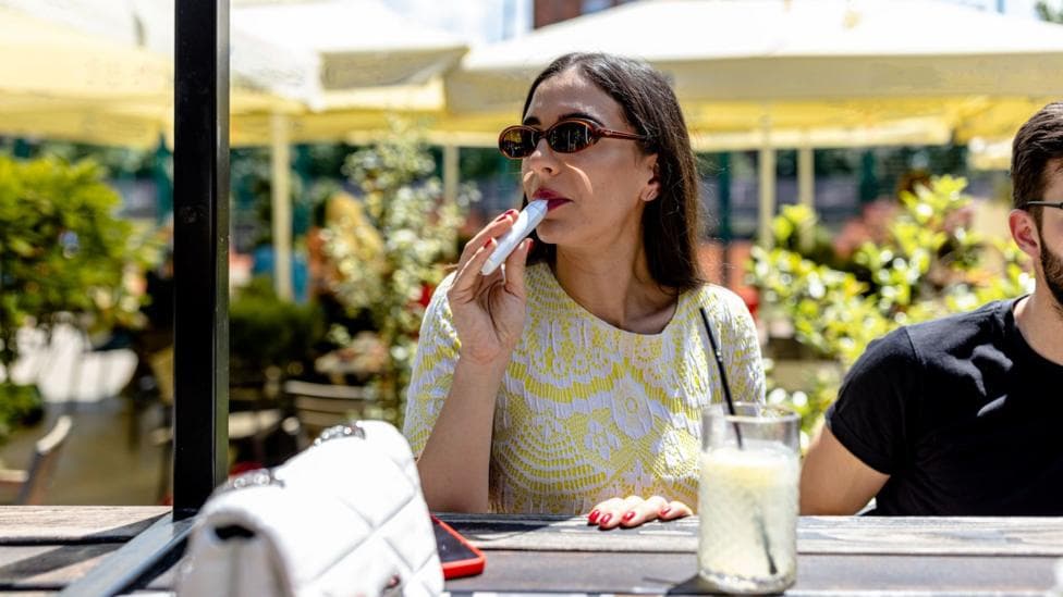 A woman vapes at an outdoor bar. She wears sunglasses and sits in the sun.
